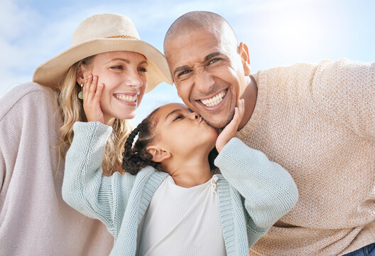 Happy family, mother and father with a kiss from a child on the face in summer outdoors on a weekend. Interracial, mama and girl loves kissing dad with a big smile on cheek on sunny holiday vacation