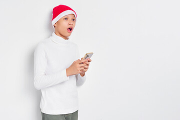 Surprised handsome Asian young man in white t-shirt and in Santa Claus hat holding mobile phone and looking aside isolated on white background. Merry Christmas and Happy New Year