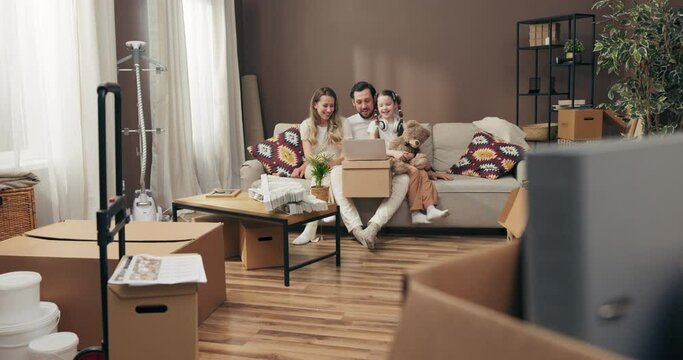 Delighted preschooler daughter with young mother and father sitting on cozy sofa in new apartment holding carton boxes full of household stuff discussing how todecorate flat. Moving in process concept