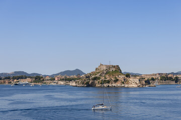 Fototapeta na wymiar Lonely small white yacht with folded sails against the backdrop of huge Old Fortress dominating all region, Corfu island, Greece, sunny morning
