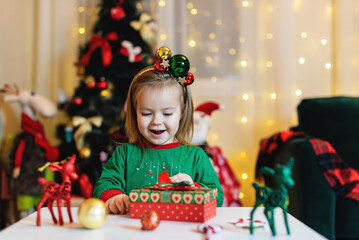Happy little smiling girl with Christmas gift box. Festive little toddler girl opening a gift at home in the living room. Christmas, holidays and childhood concept. 
