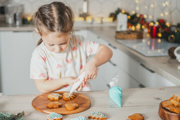 Little dark-haired girl 3 years old decorate gingerbread cookies with icing in white Christmas...