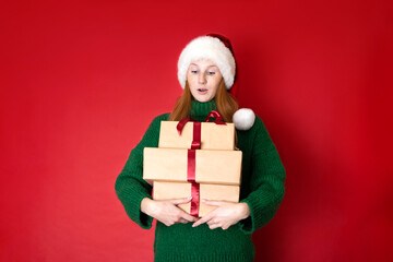 Merry Christmas Portrait of a beautiful young teenage girl in a cozy knitted green sweater and Santa's hat holding gift boxes. The red background is the place for the text.