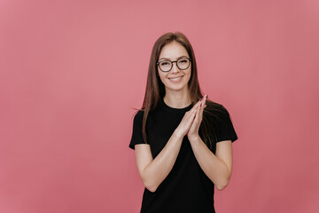 Pretty cheerful young woman in glasses with long hair in black t-shirt folding hands, with toothy smile looking at camera over pink backdrop with copy space. Grateful student, mockup. Youth, education