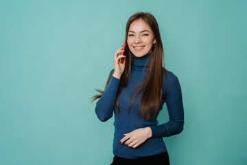 Attractive cheerful young blonde woman in blue sweater making call, using phone, looking at camera, toothy smiling over turquoise studio background with copy space. Promo, mockup of communication.