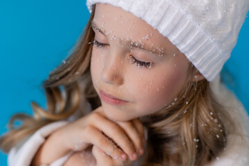 portrait of a cute little blonde girl in a white knitted white hat and sweater on a blue background in the studio. snowflakes in the hair, the concept of winter