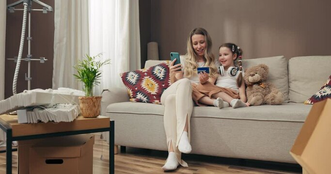 Happy young mother and preschooler kid sitting on comfortable sofa in modern new spacious apartment moving in process. Young woman holding cellphone smartphone and credit in hands typing number of