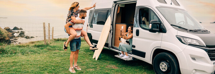 Young woman taking a photo with her cell phone of her friends having fun piggybacking next to their camper van during a trip