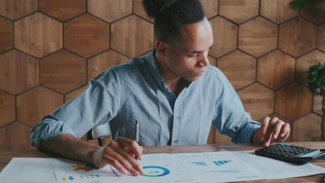 young focused African American man accountant uses calculator to draw up business plan or calculate company potential return on investment in marketing sits at table in office. Audit management