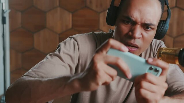 Young passionate African American man wearing headphones plays video games on mobile phone using accelerometer technology dressed in casual t-shirt sits at table with microphone. Gamer, blogger