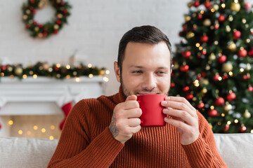 joyful man drinking warm cocoa in blurred living room with christmas decor