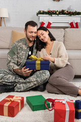 smiling woman opening christmas present while sitting on floor near husband in camouflage