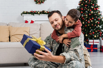 kid embracing father in camouflage holding gift box in living room with christmas decor