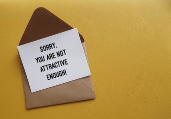 On copy space yellow background, envelope with note written SORRY, YOU ARE NOT ATTRACTIVE ENOUGH -...