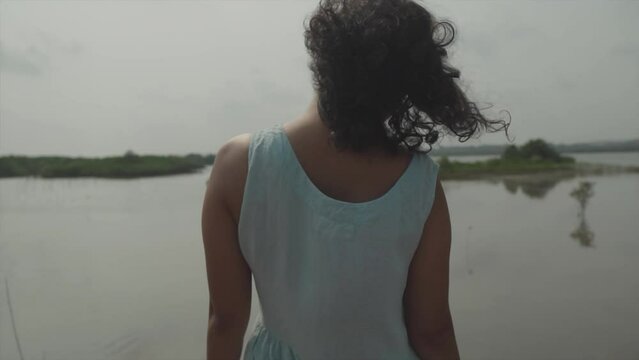 Slow motion handheld shot of a young woman from behind in light blue dress and black hair looking over a lake with small islands with plants in the distance 