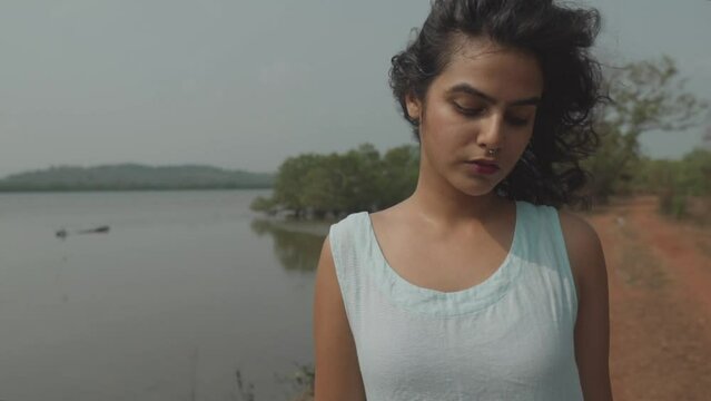 Slow motion dolly shot of a beautiful indian woman with a gold nose ring and dressed in a baby blue top standing in front of a lake with pensive look on the ground