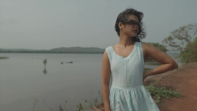 Slow motion panning shot of an attractive young woman dressed in a baby blue dress standing in front of a lake during windy weather
