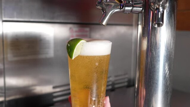 Bartender fills pint glass dressed with lime wedge with foamy draft beer from stainless keg, slow motion 4K