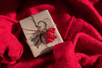 A gift in craft packaging with a sprig of viburnum against the background of a red sweater. Holiday gift. Christmas. New Year. Birthday. Valentine's Day. Holiday postcard.