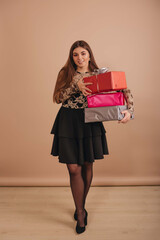 A girl with long hair, in an elegant black dress, holding many colorful gift boxes in her hands against  beige background.