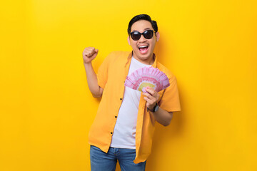 Fototapeta na wymiar Excited young handsome Asian man holding money banknotes and celebrating success isolated on yellow background