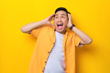 Naklejka premium Surprised young handsome Asian man holding hands on heads, looking away with open mouth isolated on yellow background