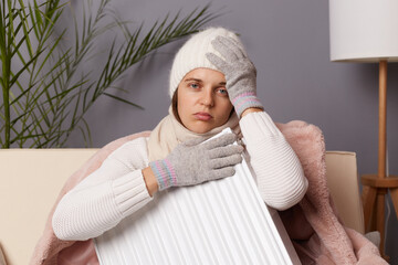 Portrait of sick unhealthy woman in coat and hat sit in cold living room with radiator, has no heating in her house, suffering flu and headache, looking at camera with sad expression.