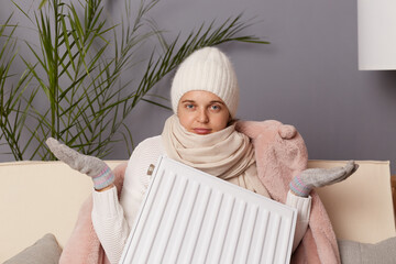Portrait of uncertain woman in coat and hat sit in cold living room holding in hand radiator,...