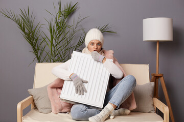 Cold frozen woman in coat and hat sit in cold living room suffering from lack of heat, blowing on...
