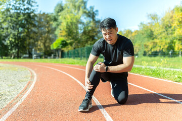 Asian man after fitness workout and jogging sits on the ground and suffers from leg pain, massages leg muscles