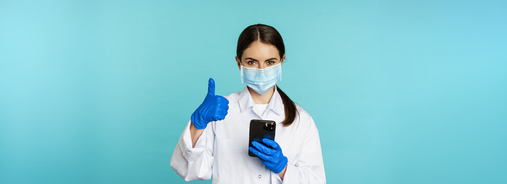 Online Doctor And Clinic. Young Woman In Medical Face Mask, Using Smartphone For Client Remote Online Appointment, Showing Thumbs Up, Standing Over Blue Background