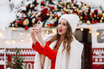 A beautiful smiling young woman millennial influencer in a fashionable knitted hat and fur coat looks into a mobile phone while standing near a decorated van on a Christmas holiday on street.  - obrazy, fototapety, plakaty