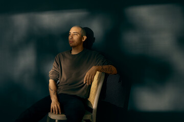 A caucasian man with a shaved head sitting against a wall on a chair posing in a ray of light a wall