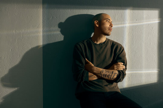 Thoughtful portrait of a pensive man sitting with crossed arms against a textured wall. Warm sunset light creates a dramatic shadow and highlights his tattoo. Cinematic atmosphere.