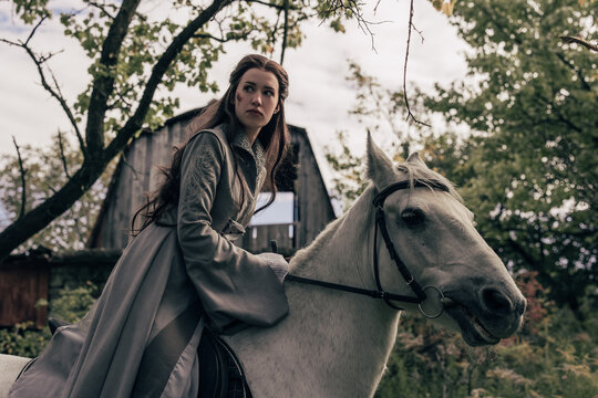 Young woman in image of ancient rider warrior sits on white horse in cloak.