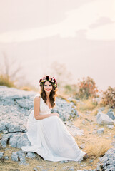 Fototapeta na wymiar Smiling bride in a flower wreath sits on stones on top of a mountain