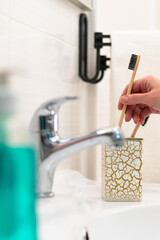 A woman's hand puts a wooden toothbrush into a glass with a gold pattern in the bathroom against the backdrop of a faucet and glossy tiles. Selective focus. Close-up