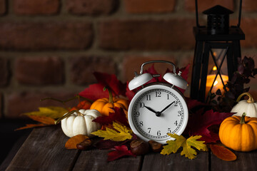 Fall winter time change concept. Autumn composition with retro alarm clock, bright yellow and red leaves, pumpkin, walnut, candle lantern. Brick wall on background. Cozy home atmosphere. Close-up