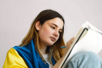 A Ukrainian teenage girl is wrapped in the Ukrainian flag and is drawing or doing homework in the bedroom. A teenager studies at home. The concept of studies during the war in Ukraine.