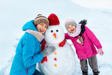 Mom and daughter are making a snowman, smiling broadly and decorating it.