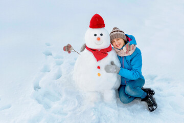 The happy girl next to a big snowman. and she decorate it with a wide smile and a warm knitted hat and scarf