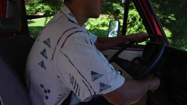 Young man sitting on driver seat is starting engine on a red retro bus, van. Green forest in the background on a sunny summer day.