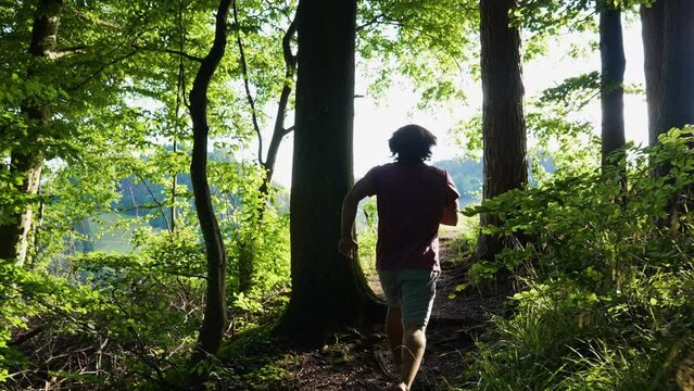 Young man with red tshirt is running barefoot in slow motion out of a dark forest towards a bright and sunny meadow. camera is following him, first person view modus.