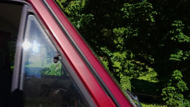 Young man gets in beatiful red retro bus, van and putting on seat belt on an sunny summer day. Green forest in background.