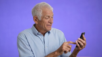 Happy fun elderly man 70s in blue shirt gets video call using mobile cell phone talk wave with hand isolated on plain light purple background studio portrait 