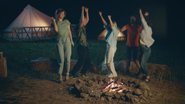 In the night time group of multiracial friends students spending time together at the campsite beside the fire stake they dancing and singing and feeling relaxed
