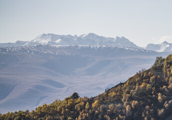 Mountain slope with trees yellowed in autumn against the backdrop of the Caucasus mountain range, warm autumn morning