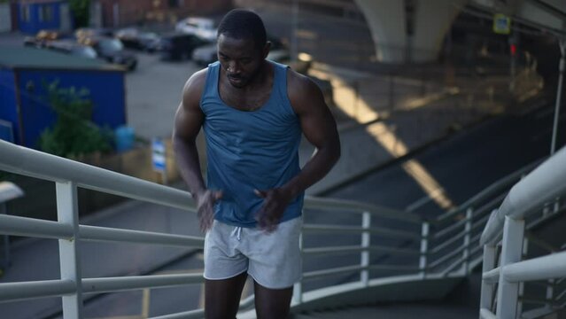 4k Male athlete runner runs up stairs in city spbas. Young american african man climbing up and doing effort exercise, posing and training outdoors in summer. Handsome muscular guy working out and