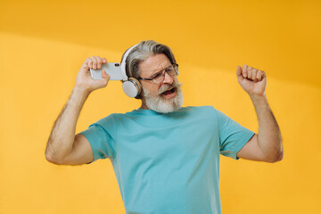 Fototapeta na wymiar Optimistic gray-haired old man listens to music, dances, wears headphones, glasses, isolated on yellow background.