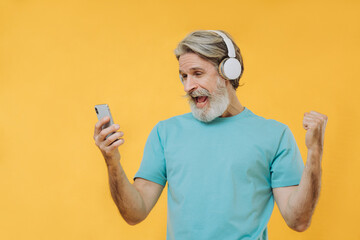 Photo of an expressive gray-haired senior man in headphones with a phone in his hands, isolated on a yellow background.